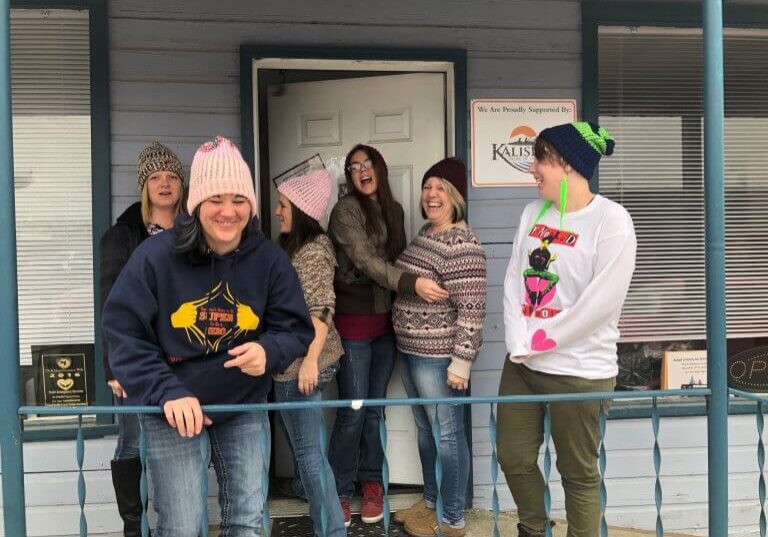 Six women wearing winter clothes stand and laugh together on a porch in front of a door, sharing a joyful moment in Pend Oreille County. A sign and plaques are visible on the building behind them.