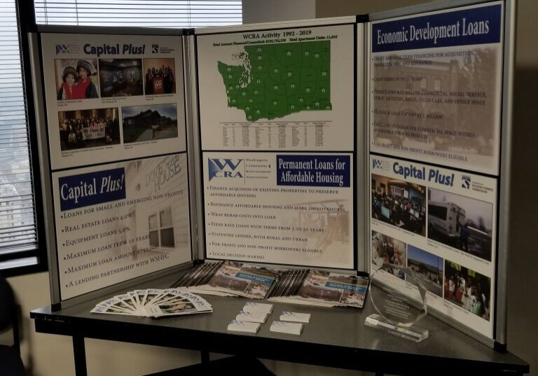 Display board for WCRA, featured at the WCRA Annual Meeting, showing information on loans for small businesses, real estate, and affordable housing, with brochures, a map, and an award arranged on a table in an office setting.