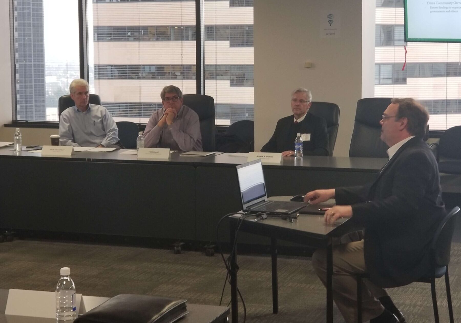 Four men in business attire are seated in a conference room during the WCRA Annual Meeting; one is presenting with a laptop while three listen attentively across the table.