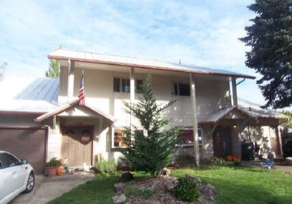 Two-story beige house with a metal roof, a covered entry, an American flag, a small front yard with plants, and a white car parked in the driveway.