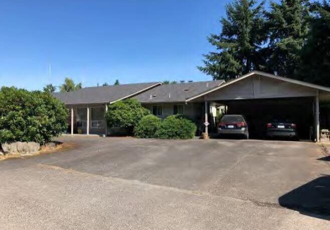 Single-story house with a carport, two parked cars, bushes in front, and tall trees in the background under a clear blue sky—an inviting scene in Valleybrook Village.