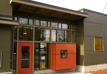 Modern building exterior with olive green walls, large windows, and an orange door and accent panel—perfectly suited for an Early Learning Center. Fallen leaves are scattered on the sidewalk in front, creating a welcoming atmosphere for families at Denise Louie.