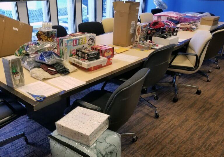 Conference room with a long table displaying board games, gifts, and packages—a festive scene that embodies the spirit of giving back during the holidays. Office chairs surround the table; papers and scissors are also visible on the tabletop, hinting at holiday charity preparations in full swing.