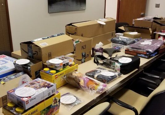 A conference table covered with assorted boxed items, toys, canned goods, and packages—all labeled with white papers—awaits distribution as part of the Mary’s Place Holiday Giving Tree initiative.