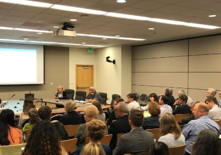 People sit in rows facing a Public Funders Panel of speakers in a conference room, with an HDC-projected presentation on public funding visible on the wall at the front of the room.