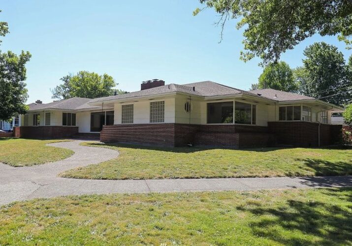 Single-story, mid-century house with brick and light exterior, large windows, and curved concrete walkways set on a grassy yard with trees in the background—an inviting example of thoughtfully designed Vets Housing.