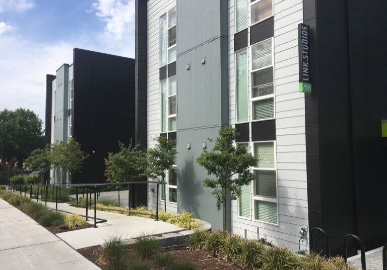 Modern apartment building, Link Studios Apartments, features a gray, black, and white exterior with large windows, small trees, sidewalk, and landscaped greenery under a partly cloudy sky.