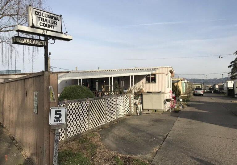 Entrance to Columbia Court Trailer Park features a 5 MPH speed limit sign, a vacancy sign, and several mobile homes lining the paved road.