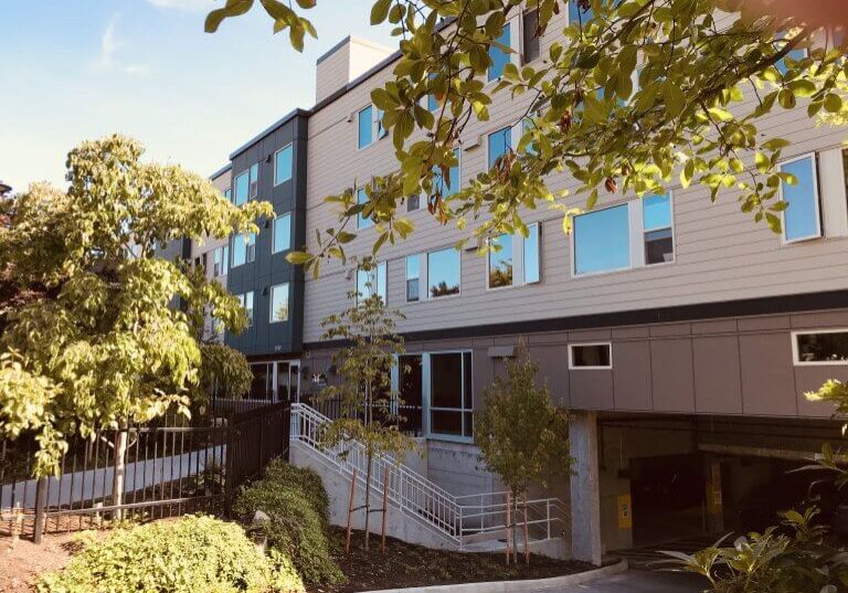 Four-story modern apartment building, 30Bellevue, features large windows and is surrounded by trees, shrubs, and meticulous landscaping; a driveway leads to an underground parking entrance, offering residents a welcoming approach to these exceptional Bellevue Apartments.