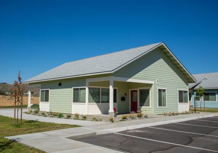 Single-story light green house with a gray roof, red front door, small garden, sidewalk, and parking spaces under a clear blue sky—perfect for comfortable family living in Meadow Point Housing.