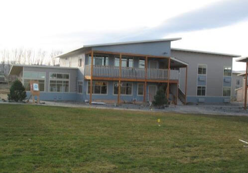 A two-story modern building with a covered balcony and large windows, surrounded by a grassy lawn and a few small trees, showcases the contemporary design elements featured in Mattawa Phase 2.