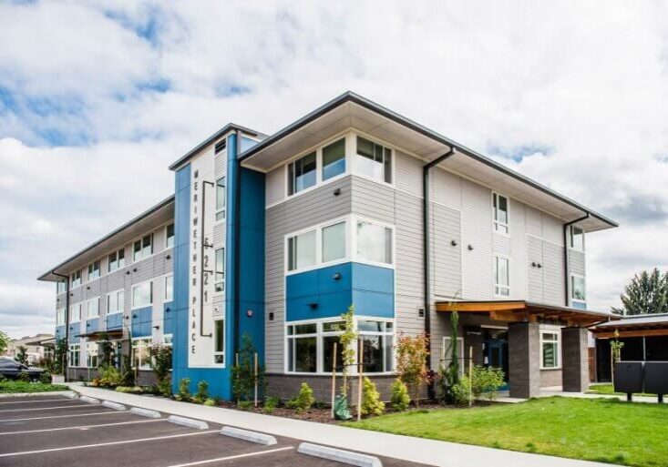 Modern three-story apartment building, Meriwether Place, features a gray and blue exterior, large windows, a landscaped entrance, and adjacent parking spaces under a partly cloudy sky.