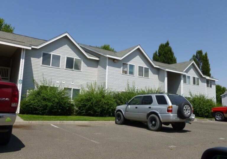 A silver SUV and a red truck are parked in front of a two-story white apartment building near Pioneer Park, with hedges and trees in the background.