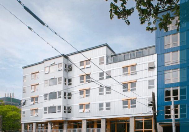 Modern five-story apartment building with gray and blue exterior panels, large windows, and yellow accents, viewed from street level with trees and power lines visible in the foreground.