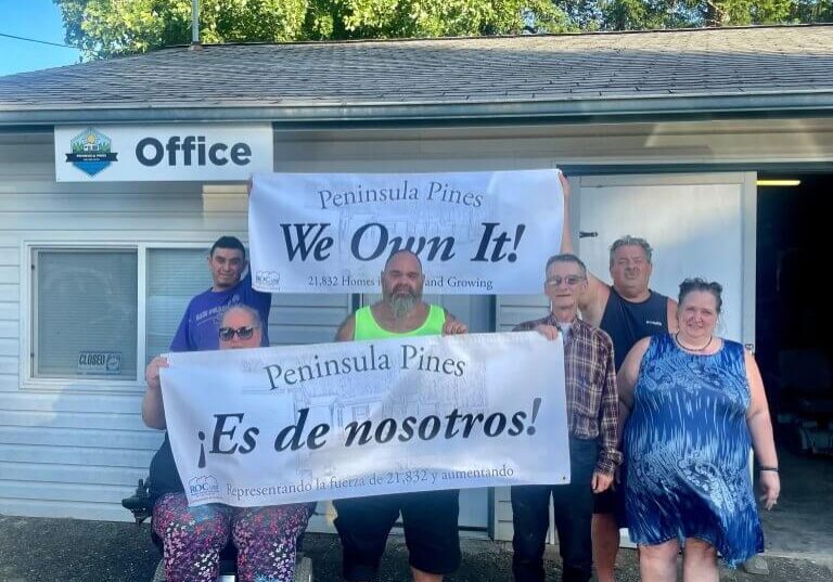 Seven people stand in front of a small building labeled “Office,” holding banners that read “We Own It!” and “¡Es de nosotros!” with “Peninsula Pines Cooperative” proudly printed on both.