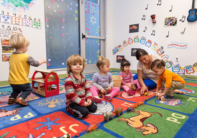 A group of young children and an adult sit and play with toys on a colorful classroom rug decorated with animals and letters.