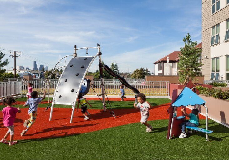 Several children play on a modern, WELL-designed playground with climbing equipment and a small playhouse, set on artificial turf beside a building and fenced area under a clear sky.