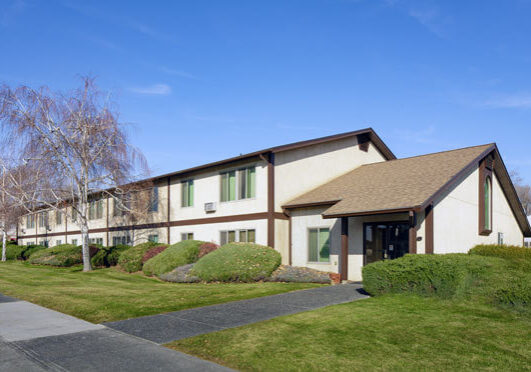 St. Martin’s Court, a two-story building with tan walls, brown trim, and green windows, sits behind bushes and a well-kept lawn under a clear blue sky.