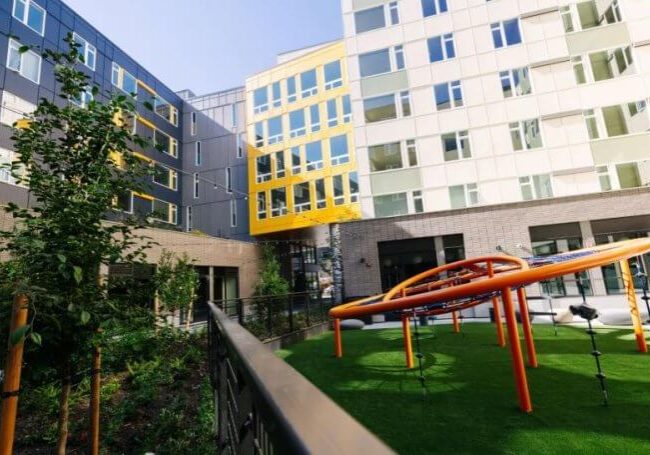 A modern apartment complex courtyard at Cedar Crossing features a small green lawn, orange playground equipment, young trees, and multi-story buildings in the background—creating an inviting space ideal for families and an inspiring Early Learning environment.