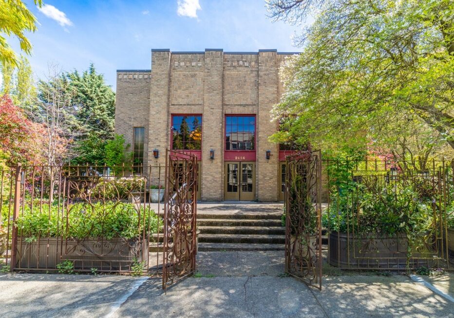 A brick building with large windows, double doors, and a decorative metal gate, surrounded by trees and greenery on a sunny day.
